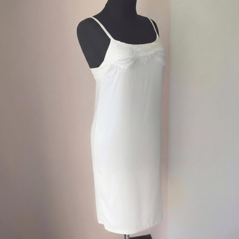 May include: A white slip dress with spaghetti straps and a lace trim detail at the neckline. The dress is displayed on a black mannequin. The slip dress is a classic piece of lingerie.
