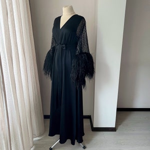 K&ouml;nnte beinhalten: Eine schwarze, lang&auml;rmelige Robe mit V-Ausschnitt und einem Bindeg&uuml;rtel. Die &Auml;rmel sind mit schwarzen Pailletten und Federn besetzt.