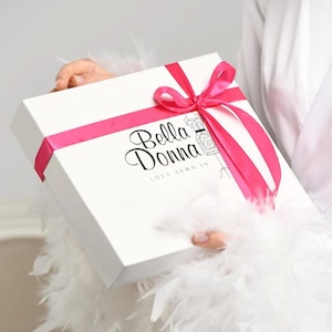 May include: A square white gift box tied with a bright pink satin ribbon. The box features the black text Bella Donna and the phrase LOVE SEWN IN, accompanied by a small line drawing of a dress form on the front surface of the packaging.