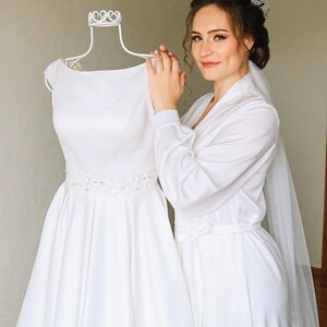 May include: A bride in a white satin robe and veil, holding a white wedding dress on a white dress form. The bride is wearing a silver tiara.