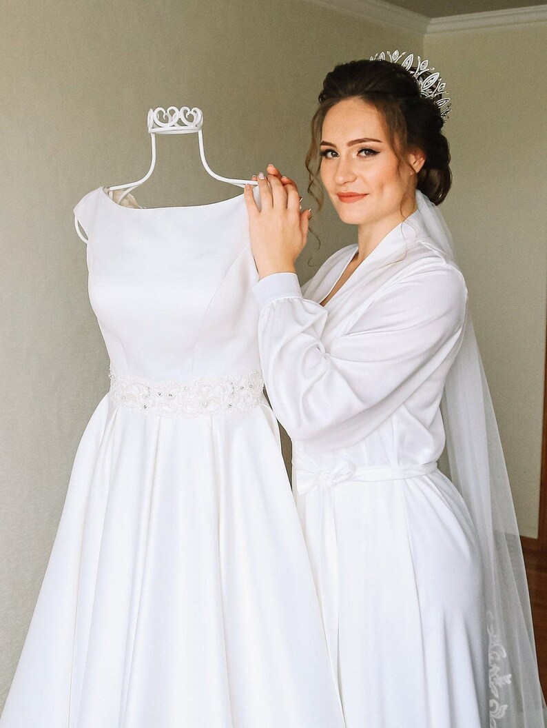 May include: A bride in a white satin robe and veil, holding a white wedding dress on a white dress form. The bride is wearing a silver tiara.