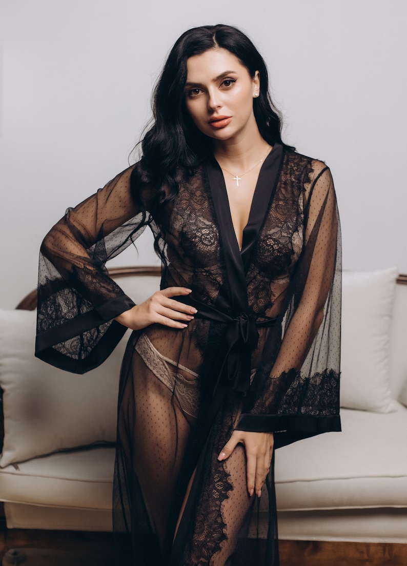 May include: A black lace robe with a satin sash and a sheer overlay. The robe has long sleeves and a V-neckline. The model is wearing a nude bra underneath the robe.