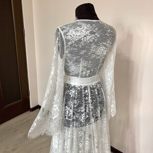 May include: A white lace dress with long, flared sleeves and a matching white belt. The dress is displayed on a mannequin, showcasing the intricate floral lace pattern. The dress has a flowing, layered skirt.