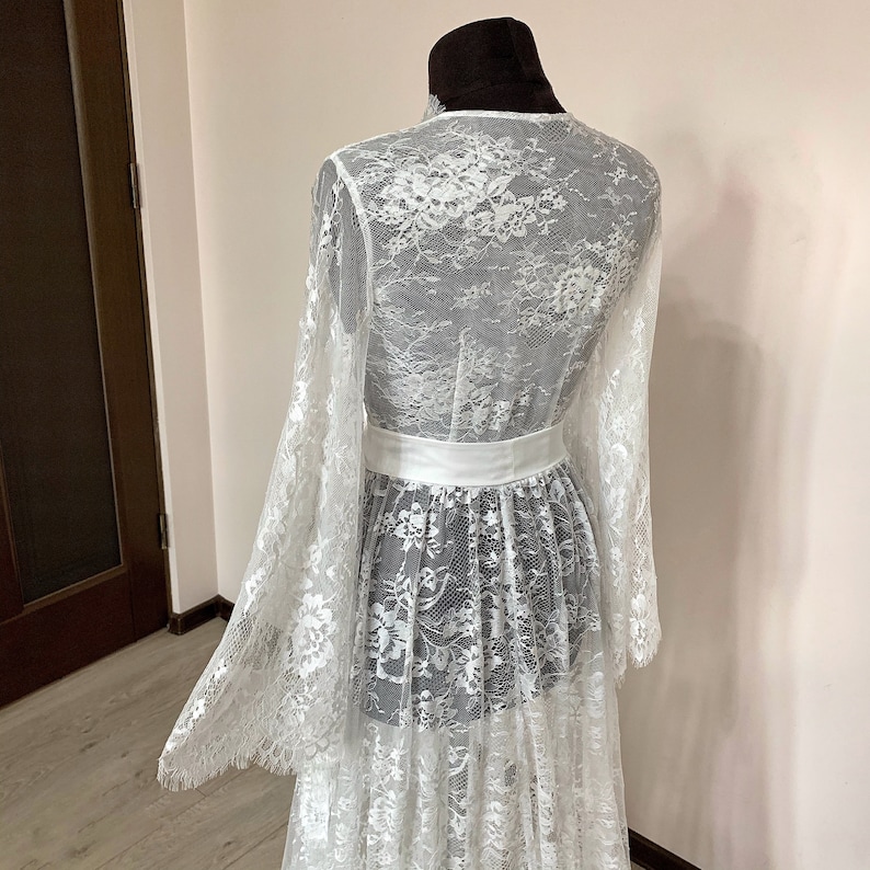 May include: A white lace dress with long, flared sleeves and a matching white belt. The dress is displayed on a mannequin, showcasing the intricate floral lace pattern. The dress has a flowing, layered skirt.