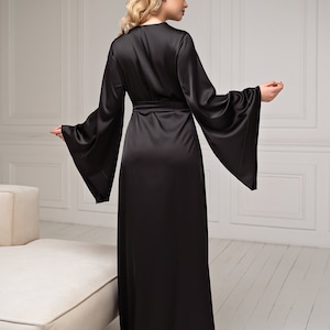 May include: A long, black satin robe with wide, flared sleeves and a matching belt. The robe is floor-length and has a wrap-around design. The model is wearing the robe in a well-lit room with a neutral background. The robe is a luxurious garment.