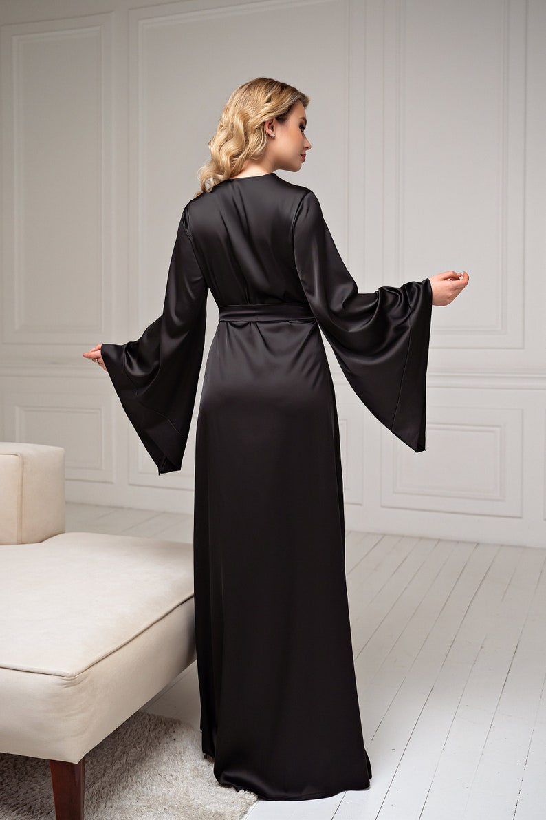 May include: A long, black satin robe with wide, flared sleeves and a matching belt. The robe is floor-length and has a wrap-around design. The model is wearing the robe in a well-lit room with a neutral background. The robe is a luxurious garment.