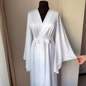May include: A white satin robe with wide sleeves and a matching tie belt. The robe has a V-neck design and is displayed on a mannequin. The sleeves are long and flared, and the fabric has a slight sheen.