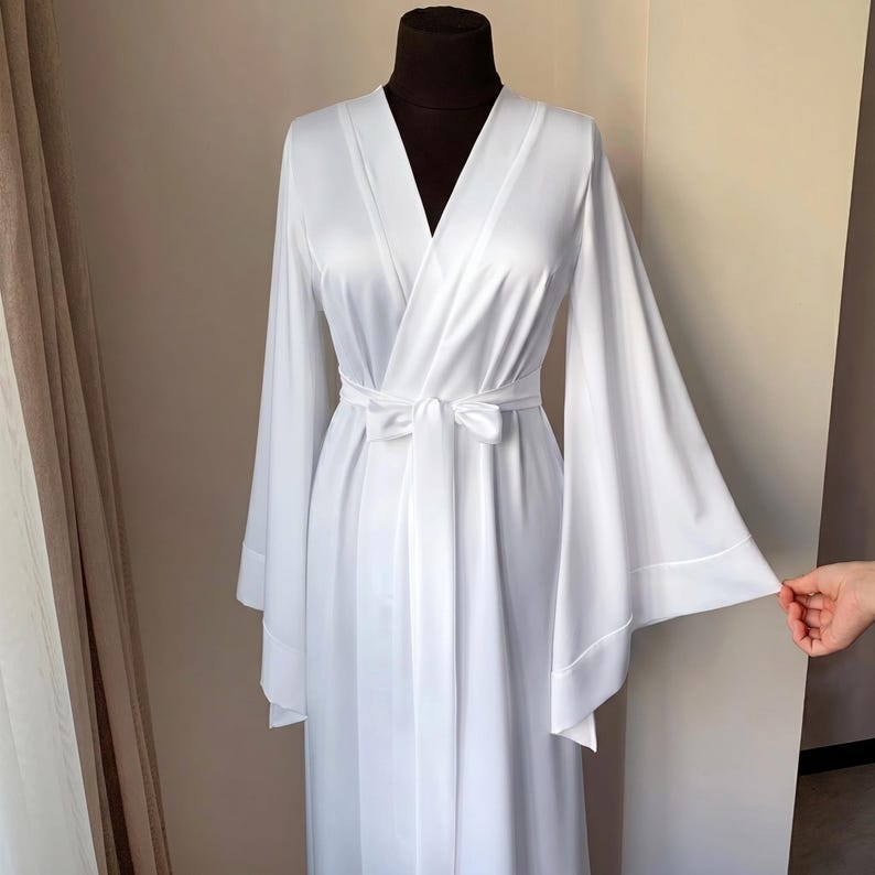 May include: A white satin robe with wide sleeves and a matching tie belt. The robe has a V-neck design and is displayed on a mannequin. The sleeves are long and flared, and the fabric has a slight sheen.