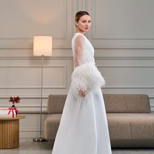 A bride stands in profile, showcasing the flowing design of a white satin bridal robe. The sheer sleeves sparkle with delicate beadwork, while the plush ostrich feather cuffs add texture and movement.