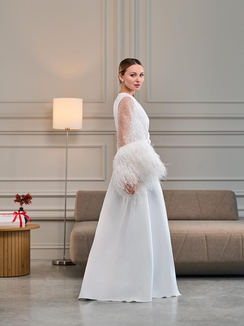 A bride stands in profile, showcasing the flowing design of a white satin bridal robe. The sheer sleeves sparkle with delicate beadwork, while the plush ostrich feather cuffs add texture and movement.