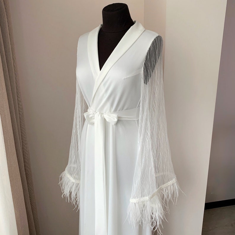 May include: A white robe featuring long, sheer, textured sleeves that flare at the wrists. The garment has a shawl collar, a matching fabric tie belt at the waist, and white feather trim detailing along the edges of the wide, flowing sleeve cuffs.