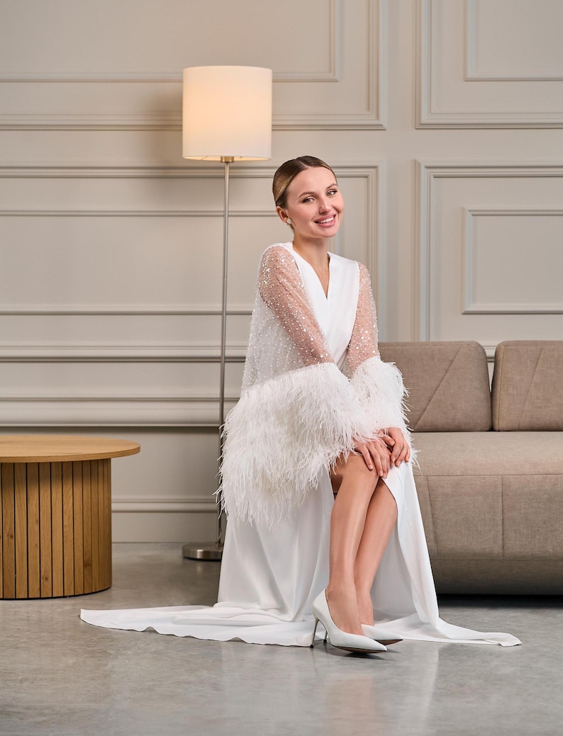 A smiling bride sits elegantly in a white silk robe with sheer beaded sleeves and feather-trimmed cuffs. The luxurious fabric cascades around her, creating a soft and romantic bridal moment.