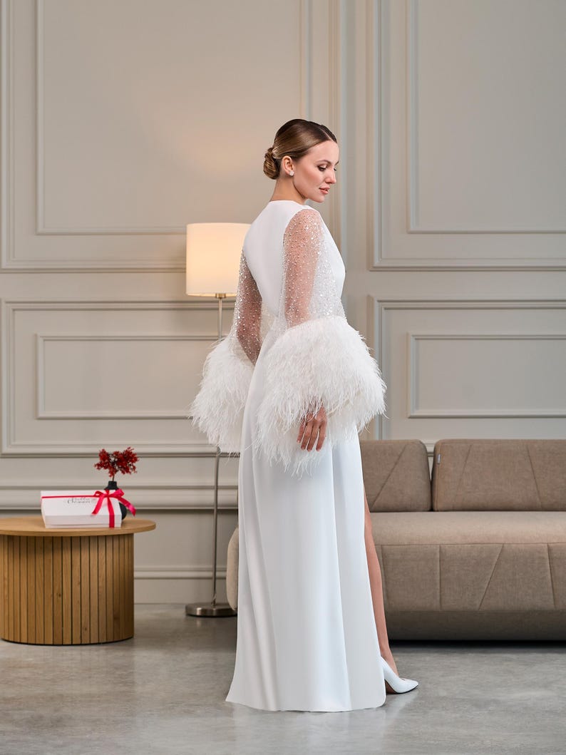 A back view of a long white satin robe with sheer pearl-embellished sleeves and voluminous feather cuffs. The smooth satin drapes gracefully, while the intricate sleeve details add a touch of vintage glamour.