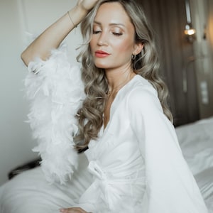 May include: A white robe featuring long sleeves with white feather trim at the cuffs. The robe has a wrap-style front with a matching fabric tie closure at the waist. The garment is made of a smooth, solid white fabric with a subtle sheen.