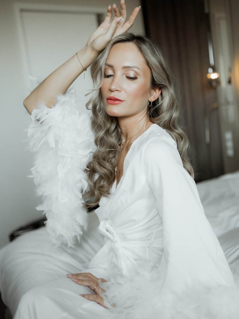 May include: A white robe featuring long sleeves with white feather trim at the cuffs. The robe has a wrap-style front with a matching fabric tie closure at the waist. The garment is made of a smooth, solid white fabric with a subtle sheen.