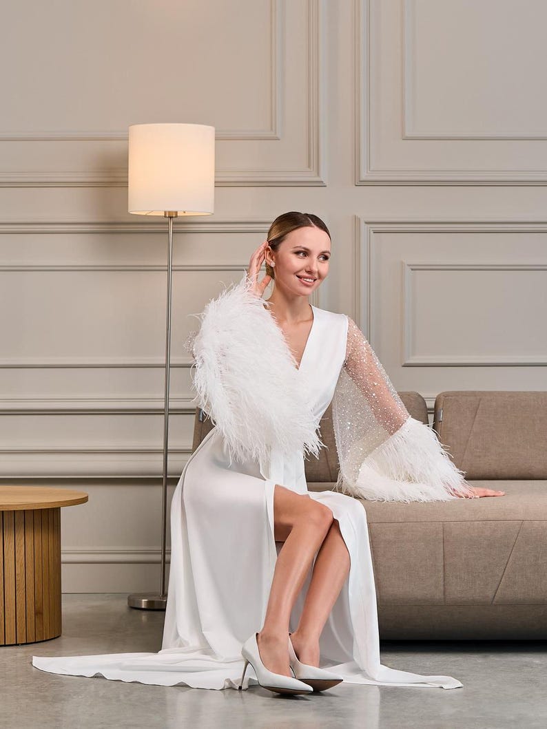 A bride lounges gracefully on a modern beige sofa, wearing a white silk bridal robe with intricate beadwork and fluffy feather cuffs. The elegant interior complements the timeless beauty of the robe.