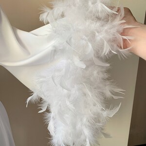 May include: A white feather boa featuring soft, fluffy plumes attached to a central cord. The long, lightweight accessory is composed of numerous delicate white feathers that create a voluminous, textured appearance throughout the entire length.