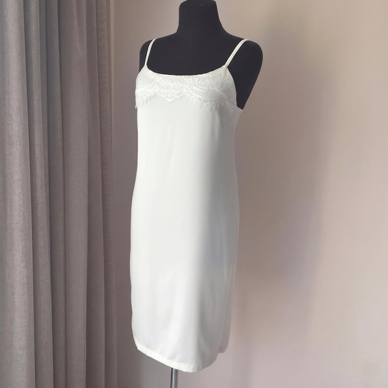 May include: A white slip dress on a black mannequin. The dress features thin spaghetti straps and a delicate lace trim along the neckline. The dress is knee-length and made of a smooth, flowing fabric.