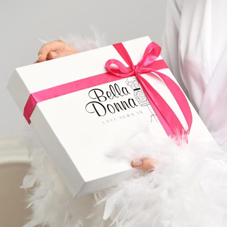 A person is holding a white gift box with a pink ribbon and the text "Bella Donna" printed on it.
