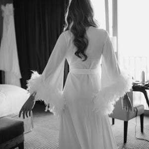 May include: A long white robe featuring wide, flowing sleeves trimmed with white feathers. The garment includes a matching fabric belt tied at the waist, creating a structured silhouette. The design is monochromatic, emphasizing the texture of the feathers.
