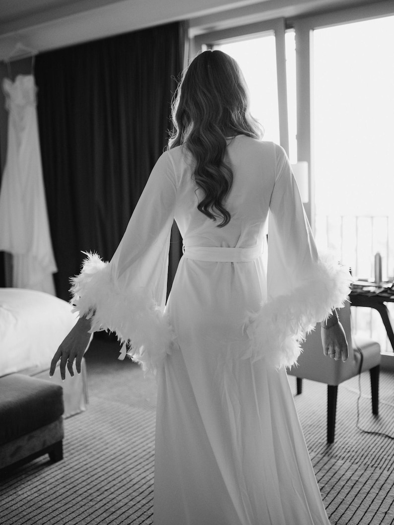 May include: A long white robe featuring wide, flowing sleeves trimmed with white feathers. The garment includes a matching fabric belt tied at the waist, creating a structured silhouette. The design is monochromatic, emphasizing the texture of the feathers.