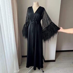 K&ouml;nnte beinhalten: Eine schwarze Satinrobe mit langen &Auml;rmeln, die mit Pailletten und Federn besetzt sind. Die Robe hat einen V-Ausschnitt und eine Bindeg&uuml;rtel an der Taille.