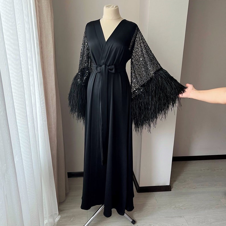K&ouml;nnte beinhalten: Eine schwarze Satinrobe mit langen &Auml;rmeln, die mit Pailletten und Federn besetzt sind. Die Robe hat einen V-Ausschnitt und eine Bindeg&uuml;rtel an der Taille.