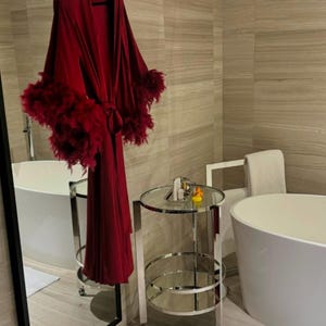 May include: A red robe with feather trim hangs on a hanger in a bathroom. The robe is tied at the waist and has long sleeves. The bathroom has a white bathtub, a silver cart with two shelves, and a mirror.