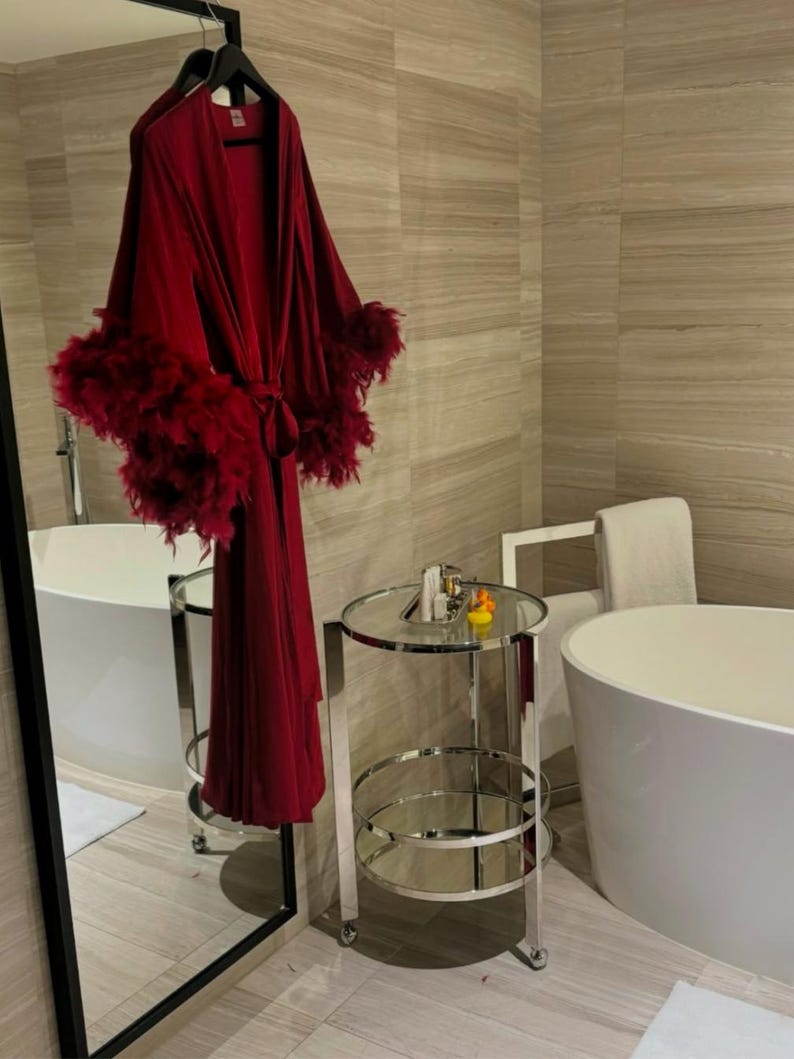 May include: A red robe with feather trim hangs on a hanger in a bathroom. The robe is tied at the waist and has long sleeves. The bathroom has a white bathtub, a silver cart with two shelves, and a mirror.