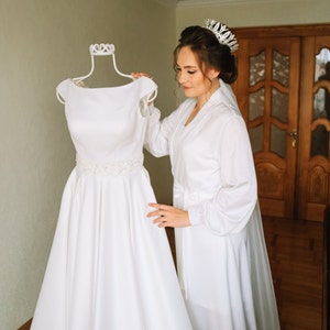 May include: A bride in a white robe and tiara is looking at a white wedding dress on a mannequin.