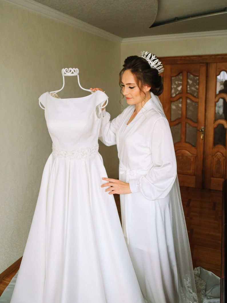 May include: A bride in a white robe and tiara is looking at a white wedding dress on a mannequin.