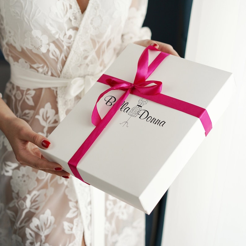 K&ouml;nnte beinhalten: Eine wei&szlig;e Geschenkbox mit einem rosa Band und dem Text "Bella Donna" auf der Vorderseite. Die Box ist mit einem rosa Band und einer Schleife gebunden.