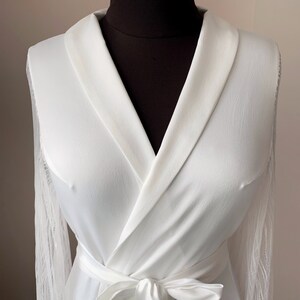May include: A white robe featuring a shawl collar, long sleeves with sheer lace detailing, and a matching fabric tie belt at the waist. The garment is displayed on a dark mannequin, highlighting the smooth texture of the main fabric.