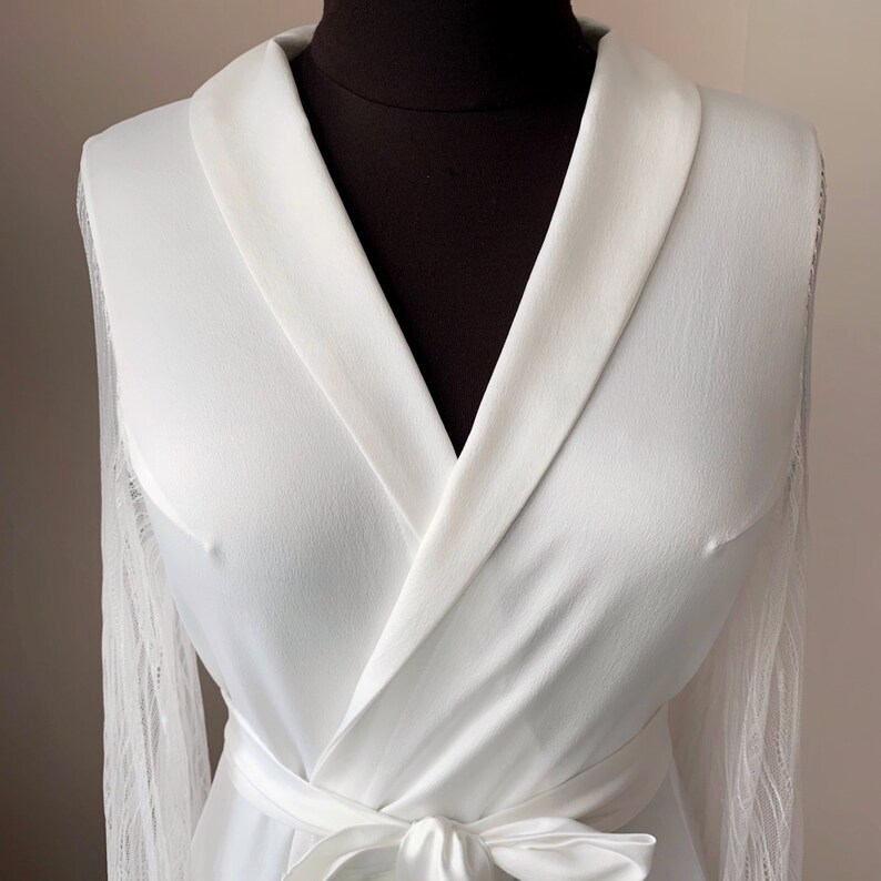 May include: A white robe featuring a shawl collar, long sleeves with sheer lace detailing, and a matching fabric tie belt at the waist. The garment is displayed on a dark mannequin, highlighting the smooth texture of the main fabric.