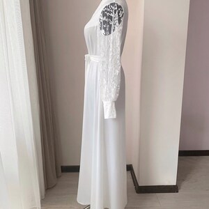 Puede incluir: Un vestido blanco largo hasta el suelo con mangas largas y transparentes que presentan un intrincado bordado floral blanco. La prenda incluye un cuello alto, un corpi&ntilde;o ajustado con un cintur&oacute;n a juego y una falda fluida.