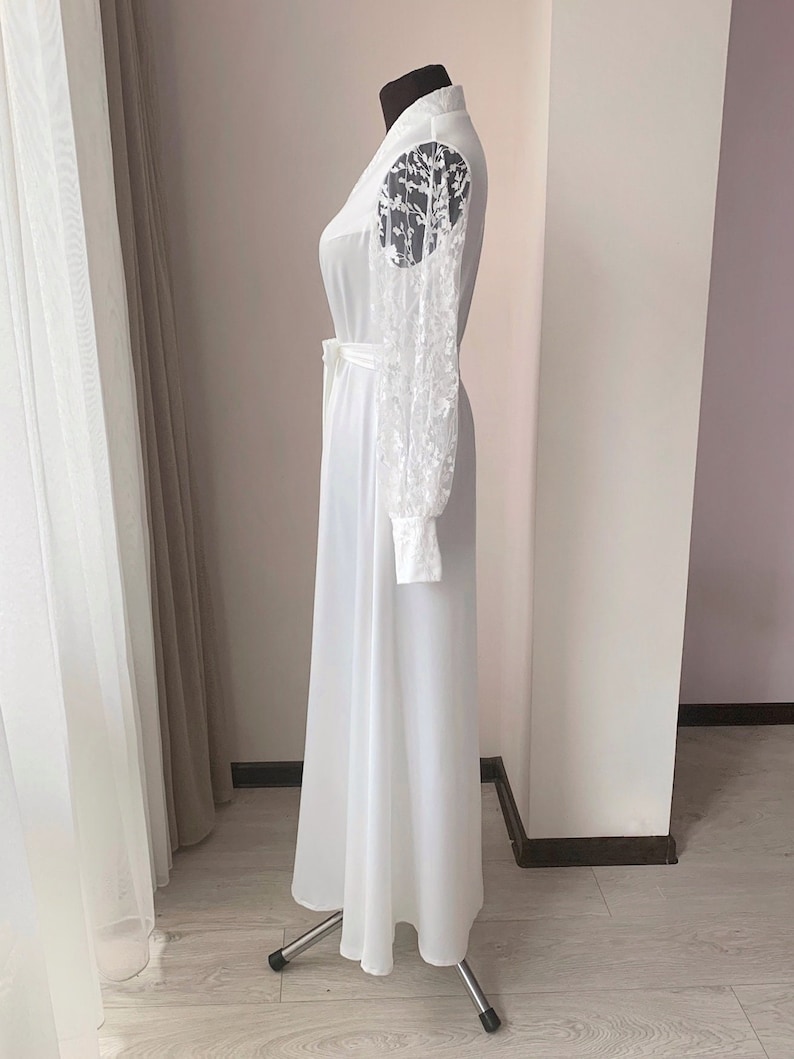 Puede incluir: Un vestido blanco largo hasta el suelo con mangas largas y transparentes que presentan un intrincado bordado floral blanco. La prenda incluye un cuello alto, un corpi&ntilde;o ajustado con un cintur&oacute;n a juego y una falda fluida.