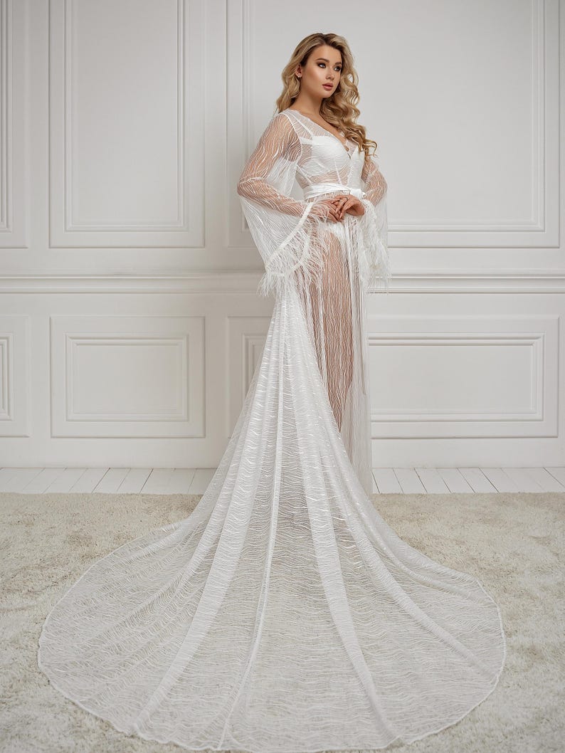 May include: A white sheer robe with a long train and feather trim. The robe has a V-neckline and long sleeves. The model is wearing a white bra underneath the robe.