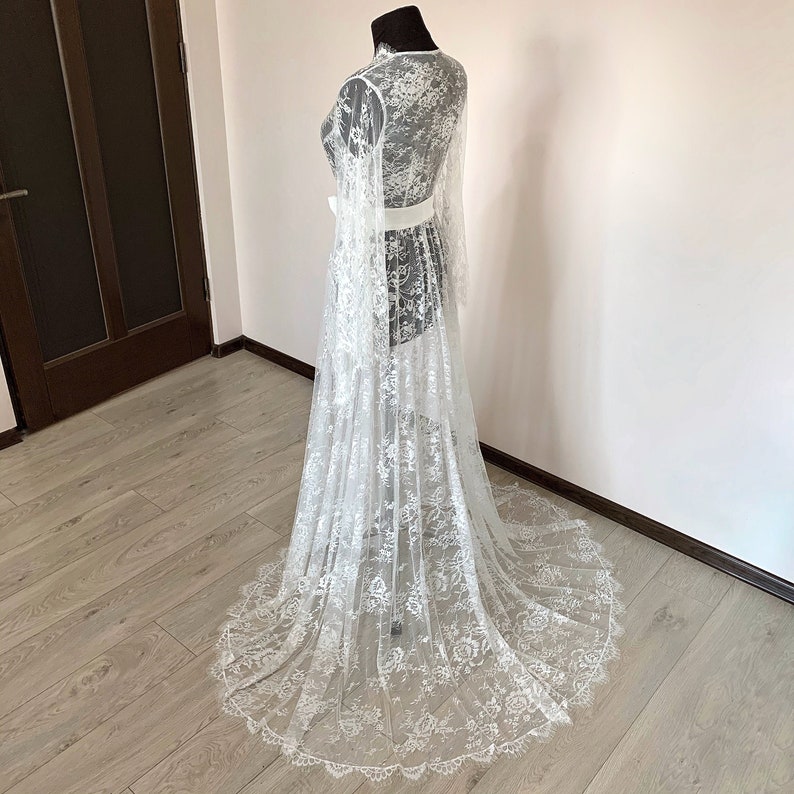May include: A white lace robe with long sleeves and a floor-length skirt is displayed on a mannequin. The robe features a high neckline, a white belt, and intricate floral lace patterns. The garment is designed for a delicate and elegant look.