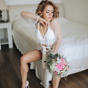 K&ouml;nnte beinhalten: Eine Frau in einem wei&szlig;en Spitzen-Dessous-Set und einem durchsichtigen Morgenmantel mit Perlenbesatz h&auml;lt einen Strau&szlig; aus rosa und wei&szlig;en Blumen. Sie tr&auml;gt wei&szlig;e Schuhe mit Absatz und sitzt am Rand eines Bettes.