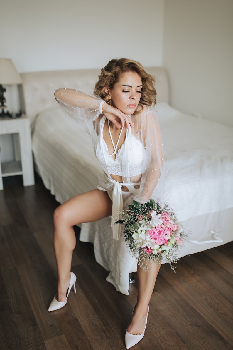 K&ouml;nnte beinhalten: Eine Frau in einem wei&szlig;en Spitzen-Dessous-Set und einem durchsichtigen Morgenmantel mit Perlenbesatz h&auml;lt einen Strau&szlig; aus rosa und wei&szlig;en Blumen. Sie tr&auml;gt wei&szlig;e Schuhe mit Absatz und sitzt am Rand eines Bettes.