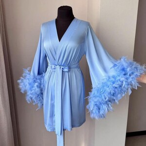 May include: A light blue satin robe featuring a V-neckline, long wide sleeves, and a matching tie belt at the waist. The cuffs of the sleeves are trimmed with thick, matching light blue feathers, creating a textured and layered appearance.