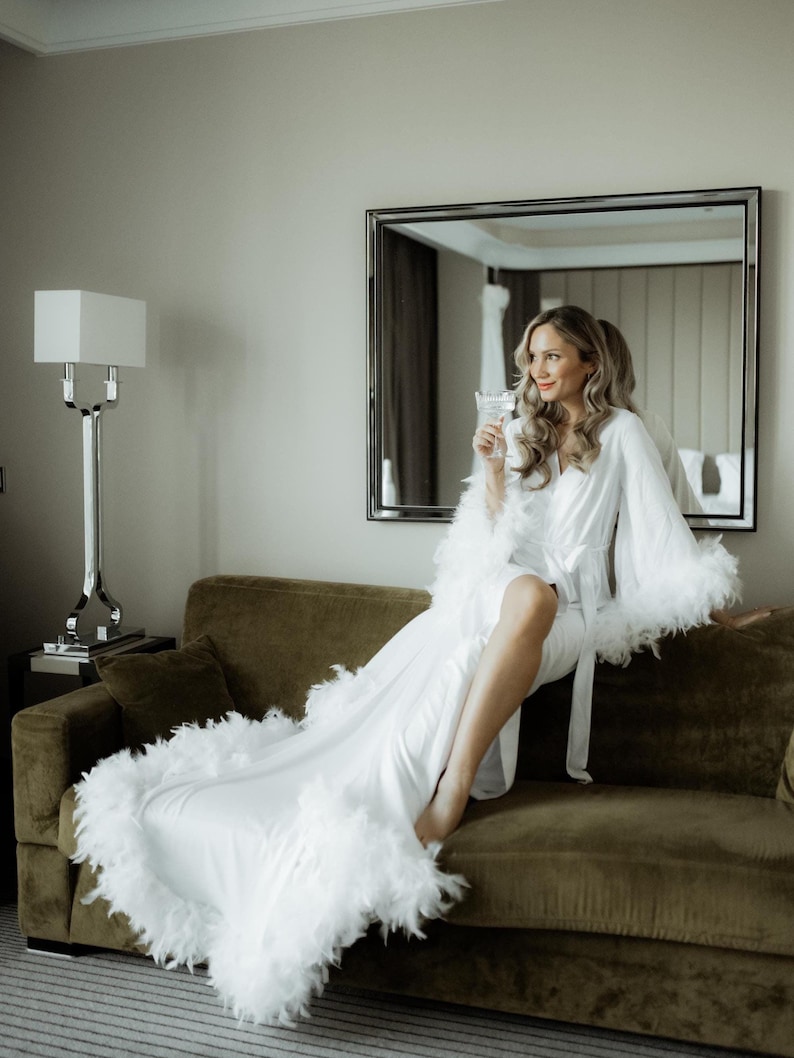 May include: A floor-length white robe featuring long sleeves and a hemline trimmed with white feathers. The robe includes a matching fabric tie belt at the waist, creating a wrap-style silhouette. The garment is made of a smooth, solid white fabric.