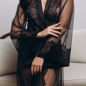 May include: A black sheer robe with lace detailing and a satin trim. The robe is open in the front, revealing a black lace bra underneath.
