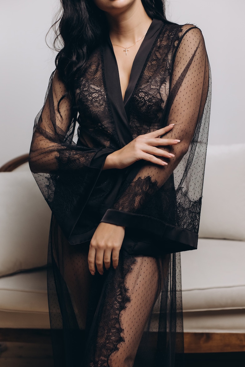 May include: A black sheer robe with lace detailing and a satin trim. The robe is open in the front, revealing a black lace bra underneath.