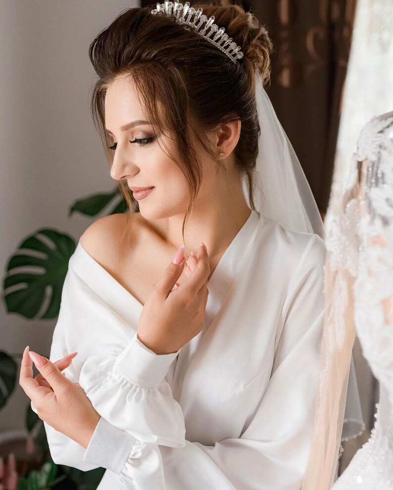 May include: A bride wearing a white satin robe with a veil and a tiara. The robe has long sleeves with ruched detailing at the cuffs.