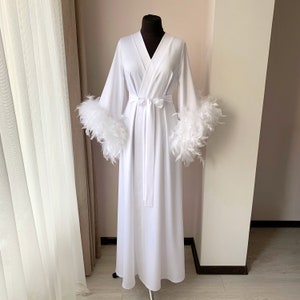 May include: A floor-length white robe featuring long, wide sleeves trimmed with voluminous white feathers. The garment has a wrap-style front with a matching fabric tie belt at the waist, creating a clean, solid white silhouette on a mannequin.