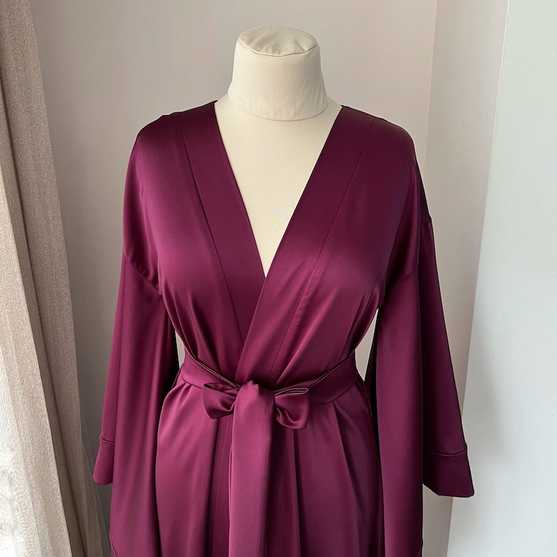 May include: A deep purple satin robe with a V-neck and long sleeves. The robe has a tie belt and a relaxed fit.