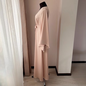 May include: A long, flowing, blush-colored robe with wide, bell sleeves and a sash tie at the waist.