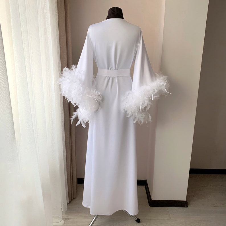 May include: A floor-length white robe featuring long, wide sleeves trimmed with thick white feathers. The garment includes a matching white fabric belt cinched at the waist, creating a structured silhouette. The design is plain, solid white.