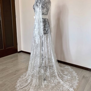 May include: A white, floor-length lace robe with long sleeves and a flowing train. The robe is cinched at the waist with a white sash. The intricate floral lace pattern covers the entire garment, creating a delicate and elegant look.